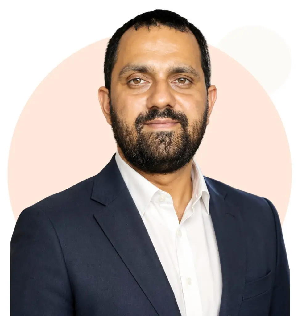 Professional headshot of a man with a beard wearing a dark blue suit jacket and white shirt, looking directly at the camera against a light beige and gray background.